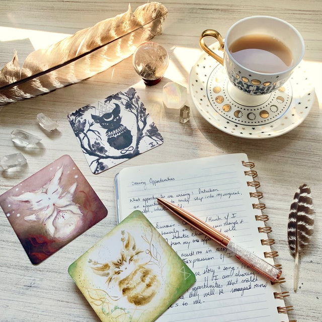 A table layout with a diary, pen, feather, crystals, teacup, and three cards from the Spirit Cats Oracle Deck