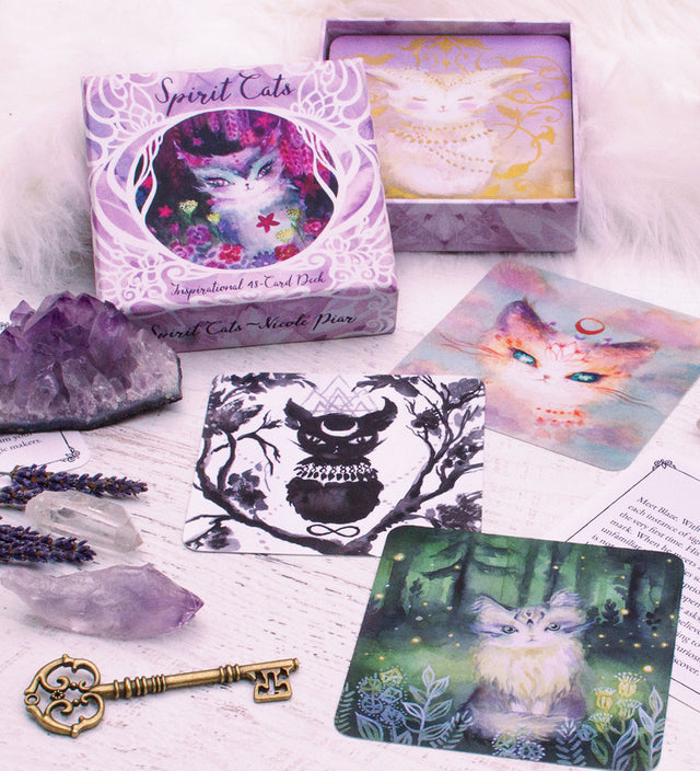 A selection of purple colored cards from the Spirit Cats Oracle deck with a deck box, key, and amethyst stone around them.