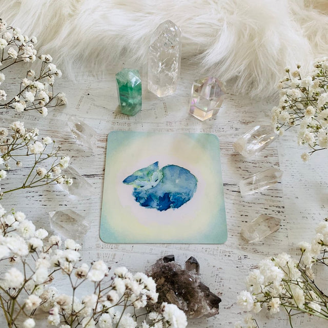A card from the Spirit Cats Oracle Deck with a turquoise kitten sleeping, and crystals and baby's breath flowers around it.