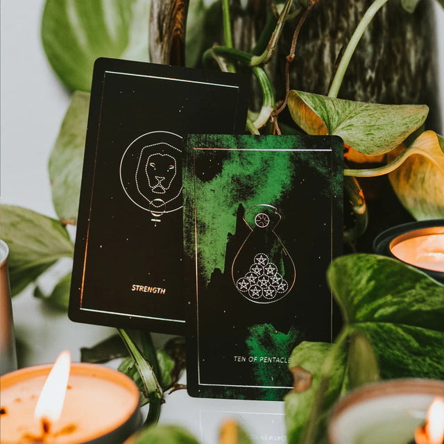 Two green cards - Strength and the Ten of Pentacles - from the Synesthesia Tarot deck with a candle and greenery around them.